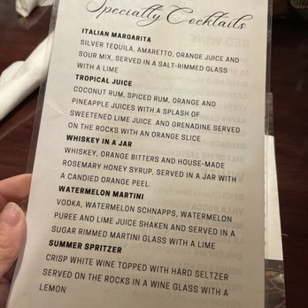 Primo - Cocktail menu - A menu titled "Specialty Cocktails" lists various drink options including Italian Margarita, Tropical Juice, Whiskey in a Jar, Watermelon Martini, and Summer Spritzer. The menu describes ingredients and presentation details for each cocktail. It is held by a person in a restaurant setting, with a hand visible holding the menu and a blurred background showing a person with folded hands and a table. - Italian Near Me - New Castle, Indiana