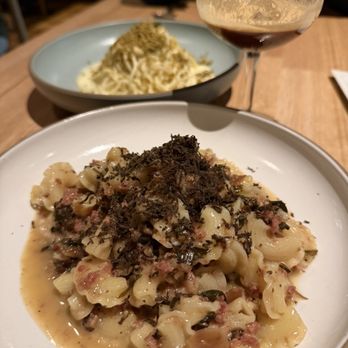 Tavernetta - The image shows two plates of pasta on a wooden table in a restaurant. The foreground features a plate of pasta with a creamy sauce, topped with black truffle shavings. In the background, there is another plate of pasta with a different sauce, possibly with herbs or cheese. A glass of red wine is positioned behind the plates, and a bowl of rice is also visible. The setting appears to be a casual dining environment with warm lighting. - Italian Near Me - Denver, Colorado
