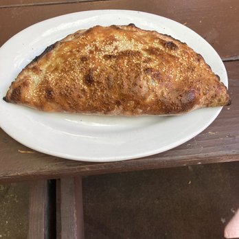 La Val's Pizza - Greek Calzone.  It's a great tasting veggie dish that you will love - A baked calzone with a crispy, golden-brown crust topped with sesame seeds is displayed on a white oval plate. The calzone appears to be filled with ingredients such as vegetables or cheese. It is positioned on a wooden table surface, suggesting a casual dining setting, possibly at a pizzeria or restaurant. - Pizza Near Me - Berkeley, California