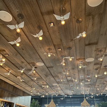 ilili DC - Ceiling - The ceiling is made of wooden planks and is decorated with numerous white bird-shaped light fixtures and exposed bulbs hanging from cords. The fixtures are arranged in a scattered pattern, providing ambient lighting. The space below includes large windows, greenery, and decorative hanging elements, suggesting a stylish interior setting. - Lebanese Near Me - Washington, DC, District of Columbia