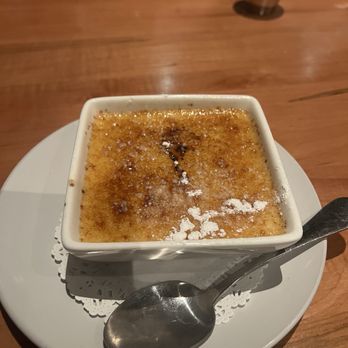 Bluestone Restaurant  - Creme Brûlée - Crème brûlée is presented in a square ramekin on a white plate, with a spoon placed beside it, on a wooden table. - American Near Me - Timonium, Maryland