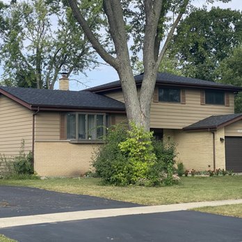 DC &T Painting - Before - A two-story residential house with beige siding, brick lower walls, and a dark garage door, situated in a suburban neighborhood. - Painters Near Me - Rolling Meadows, Illinois