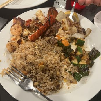 EZBachi - Lobster, steak and shrimp. - A white round plate holds fried rice with small pieces of vegetables, grilled seafood including shrimp and lobster, and stir-fried zucchini and onions. A fork is placed on the plate, and a hand is seen holding a spoon with some food. The background includes an empty plate with chopsticks and a dark table surface. - Teppanyaki Near Me - Phoenix, Arizona