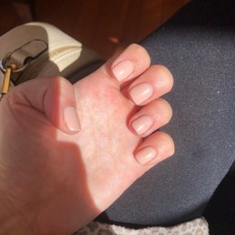 The image shows a person's left hand with well-groomed, natural-colored nails, resting on their lap. The person is wearing dark pants and a leopard-print garment. A beige purse with a zipper and gold hardware is visible in the background, placed on a wooden surface. The lighting highlights the nails and the texture of the skin.