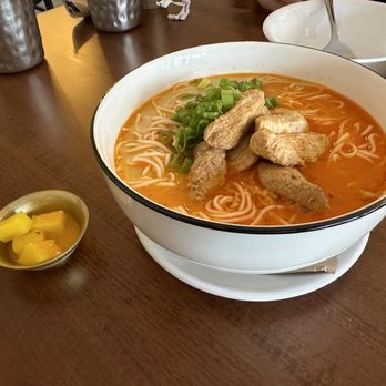 Momo Valley - A bowl of noodle soup with a red, spicy broth contains cooked noodles, tofu pieces, and chopped green onions. The bowl is placed on a wooden table, with small bowls and metal cups nearby, suggesting a restaurant setting. A small dish of pickled vegetables is also visible beside the bowl. - Himalayan/Nepalese Near Me - Beacon, New York