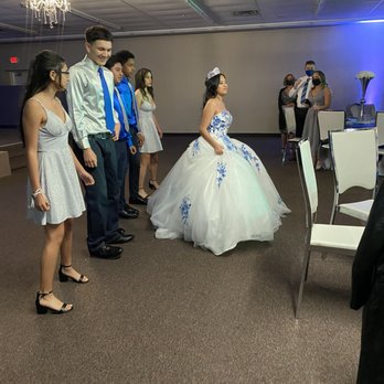 Elevate Event - A girl wearing a white gown with blue floral accents and a tiara stands in front of a group of young people at a formal event. The group includes boys and girls dressed in formal attire, with some wearing masks. The setting appears to be a banquet hall with a chandelier and decorated tables in the background. - Venues & Event Spaces Near Me - Pflugerville, Texas