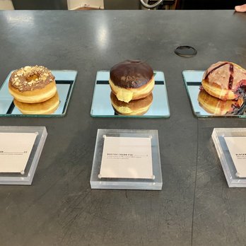 DOZER Coffee - Donut display part 3! - Three donuts are arranged on small square mirrors on a dark countertop, each with a label in front. The donuts vary in toppings and fillings, including a crumb-topped donut, a chocolate-glazed donut, and a berry-topped donut. The labels indicate the flavors: Nutella Crunch, Boston Cream Pie, and Blackberry Lemonade. The setting appears to be a bakery or café display, with a focus on presenting the donuts for tasting or sale. - Cafes Near Me - Ann Arbor, Michigan