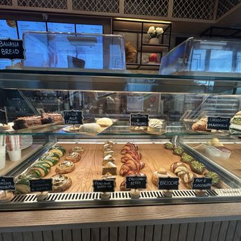 Pistacchio Pane e Caffe - Sinful Pastries to try - A bakery or cafe display case showcases an assortment of baked goods and sandwiches, including banana bread, croissants, pistachio cream rolls, and berry-filled pastries. Labels identify each item, and the case is situated in a cafe setting with a menu screen and decorative elements in the background. - Bakeries Near Me - Miami, Florida