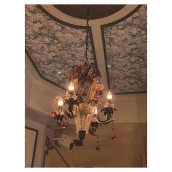 Yours Sincerely - A chandelier featuring multiple light bulbs and ornate glass decorations is suspended from a ceiling. The ceiling has a floral-patterned design, and the chandelier's chain is visible. The lighting creates a warm ambiance in the room. - Cocktail Bars Near Me - Brooklyn, New York