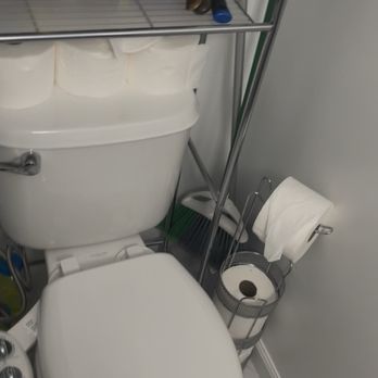 Ashbram Cleaning Services - Cleaned bathroom and organized over toilet shelving. Sparkling bathroom floor and toilet. - A small bathroom features a toilet with a storage shelf above it, holding various toiletries and a pink box. A toilet paper holder with a roll and a small trash bin are also visible. - Office Cleaning Near Me - Brambleton, Virginia