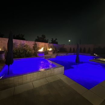 Bella Vista Pools - Spa, Baja, fire bowls, changeable lightning - Nighttime view of a backyard pool with blue LED lighting, fire bowls along the perimeter, and a surrounding brick wall with plants. - Pool & Hot Tub Service Near Me