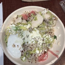 Baja bowl with 3 pieces of chicken. Not worth the money - A ceramic bowl contains a rice-based dish topped with sliced chicken, diced avocado, shredded cheese, and a poached egg. Green sauce and pickled vegetables are also present, with the dish placed on a wooden table. Napkins and a drink are visible in the background, indicating a casual dining setting.