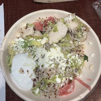 Panera Bread - Baja bowl with 3 pieces of chicken. Not worth the money - A ceramic bowl contains a rice-based dish topped with sliced chicken, diced avocado, shredded cheese, and a poached egg. Green sauce and pickled vegetables are also present, with the dish placed on a wooden table. Napkins and a drink are visible in the background, indicating a casual dining setting. - Sandwiches Near Me - Peoria, Illinois