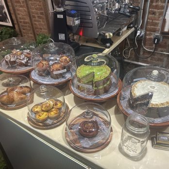 11:11 CAFE - Pastries - Multiple pastries and cakes, including croissants, tarts, muffins, and slices of cake, are displayed on a counter with glass covers. The background shows a coffee machine, cups, and a brick wall, indicating a cafe or bakery environment. - Cafes Near Me - Brooklyn, New York