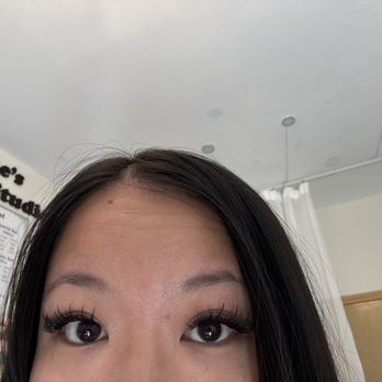 She’s Lash Studio  - Wispy volume - A woman with long eyelashes and dark hair is taking a selfie indoors, with a menu and part of a wall visible in the background. - Eyelash Service Near Me - Brooklyn, New York