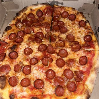 Rehoboth Baking - New Haven style pepperoni pizza - A pizza topped with pepperoni slices and melted cheese is shown inside an open cardboard pizza box. The crust has some charring around the edges, indicating it was baked at a high temperature. The pizza is cut into slices, with one slice slightly pulled away from the rest. The box is placed on a flat surface, and part of the box's interior design is visible in the background. - Bakeries Near Me - Rehoboth, Massachusetts