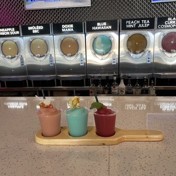 Doxie Slush - Flight (L to R): Strawberry Amaretto Shortcake, Blue Hawaiian, Strawberry Hibiscus Frose - Three frozen cocktails in small cups are placed on a wooden tray on a bar counter. The drinks are garnished with fruit and mint, with straws in each. Behind the bar, there are frozen drink machines labeled with various flavors and bottles of alcohol visible underneath. The setting appears to be a casual bar or restaurant with a focus on frozen beverages. - Seafood Near Me - Hermann, Missouri