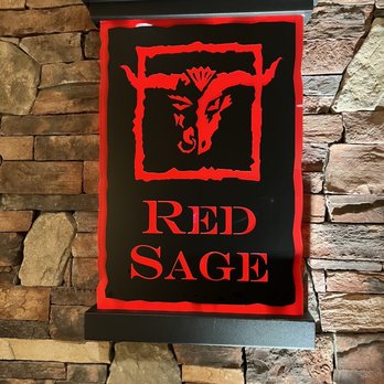 Red Sage - a sign on a brick wall - The image shows an illuminated sign for Red Sage restaurant mounted on a stone wall. The sign features a stylized red dragon logo within a red-bordered black rectangle, with the words "RED SAGE" in red capital letters below the logo. The sign has a black frame and is positioned against a textured stone wall composed of variously sized and colored stones, creating a rustic appearance. - New American Near Me - Santa Fe, New Mexico