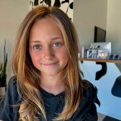 Mint Salon + Barbering - A young girl with long, honey-blonde hair and blue-green eyes smiles. Her hair features light-colored face-framing highlights...