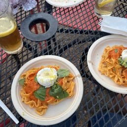 Two disposable plates of pasta with tomato sauce, basil, and poached eggs are on a black metal outdoor table, accompanied by two glasses of beer.