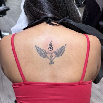 Hao Tattoo - New tattoo by Rachel at Hao Tattoo. The Khmer Om symbol. - The image shows a tattoo on the upper back of a person with long dark hair. The tattoo depicts a heart with wings, with a copyright symbol above it. The person is wearing a pink spaghetti strap top, and the background includes chairs and a person standing nearby, indicating an indoor setting such as a tattoo studio. - Tattoo Near Me - Bayside, New York