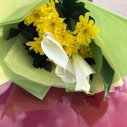 Yellow mums and white canna lillies surrounded by blackened greens wrapped in yellow and green paper with black and yellow ribbon.