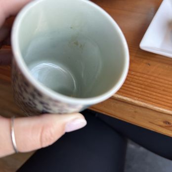 Sushi Pure - Dirty tea cup only one shown. Both were dirty - A person holds an empty ceramic mug with some stains inside, positioned over a wooden table with dishes and bowls nearby. - Sushi Bars Near Me - Eugene, Oregon