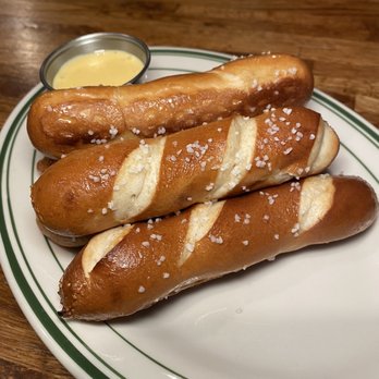 Cover Two - Pretzel sticks - Three salted pretzel sticks are arranged on a white plate with green stripes, accompanied by a small cup of mustard sauce, on a wooden table. - American Near Me - Newtown, Connecticut