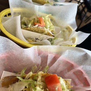 Toucan Taco - Three tacos with lettuce, tomato, and other toppings are presented in paper wrappers on colorful trays. The tacos appear to contain seasoned meat and are garnished with fresh vegetables. The setting suggests a casual dining environment, possibly at a Mexican restaurant or taqueria. - Mexican Near Me - Laurel, Maryland