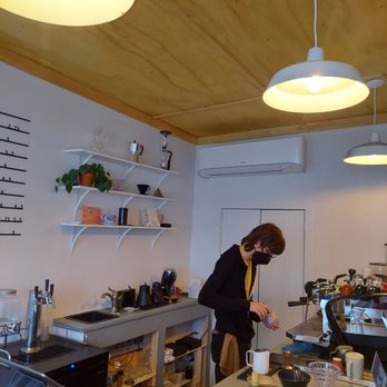 Alias Coffee - A barista, dressed in a black jacket and wearing a black face mask, is working behind a coffee shop counter. The counter has various coffee-making tools, cups, and a coffee machine. The background features white shelves with decorative items and a wall-mounted air conditioner. The ceiling has wooden panels and hanging lights, creating a cozy atmosphere. - Coffee & Tea Near Me - Troy, New York