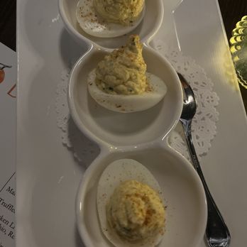 Bar Clementine - Three deviled eggs are arranged in individual small white bowls on a rectangular tray. Each egg is topped with a creamy filling, garnished with paprika, and served with a spoon. The presentation includes a doily underneath the bowls, and the tray is placed on a dark surface, possibly in a restaurant setting. - Cocktail Bars Near Me - Islip, New York