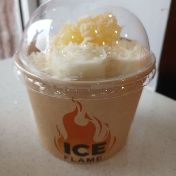 Ice Flame - Pina Colada rolled ice cream $8.49. - A plastic cup containing rolled ice cream topped with whipped cream and a drizzle of honey. The cup has a flame logo and the words 'ICE FLAME' printed on it. The ice cream appears creamy and is served with a clear plastic dome lid. The background shows a light-colored surface and part of a window or wall. - Desserts Near Me - Ottawa, Ontario