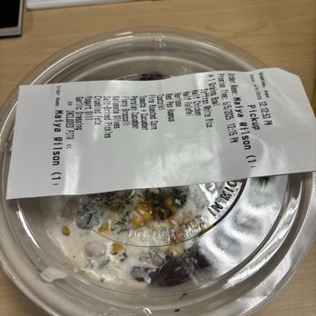 CAVA - A transparent plastic takeout container with a sealed lid is placed on a light wooden table. A receipt is laid across the top of the container, displaying order information including restaurant name, order time, and menu items. In the background, there is an open planner or calendar on the table, with visible pages and a spiral binding. - Salad Near Me - Coppell, Texas