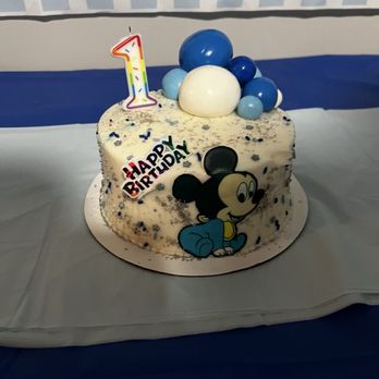 Market Basket - Cake - A birthday cake features Mickey Mouse on the side, with a 'Happy Birthday' sign and a colorful number one candle on top. The cake is decorated with blue and white balloons made of fondant or similar material, and sprinkled with small silver and blue confetti. It is placed on a white cake board, set against a background with blue and white striped decorations, indicating a birthday party setting. - Grocery Near Me - Fall River, Massachusetts
