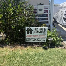 Restore & Construct - A business sign for Restore & Construct is positioned in a garden area with plants, advertising roofing, painting, windows, and remodeling services.