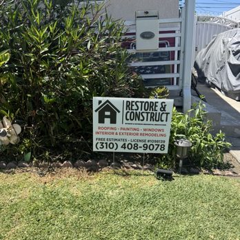 Restore & Construct - Restore & Construct - A business sign for Restore & Construct is positioned in a garden area with plants, advertising roofing, painting, windows, and remodeling services. - Roofing Near Me - Carson, California