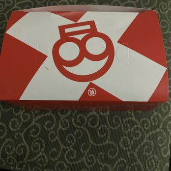 Toxic Burger - The burger and fries all fit in this box - A rectangular box with a red and white geometric design and a stylized smiley face logo on the top surface. The box is placed on a fabric surface with a swirling pattern. The logo includes circular eyes and a curved mouth, with a small registered trademark symbol below it. - Chicken Wings Near Me - Eugene, Oregon