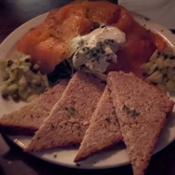 Two Fools Tavern - Salmon appetizer - A plate contains a serving of cooked salmon, three toasted bread triangles, and a side of creamy herb-infused sauce or dip. The food is arranged on a white plate, placed on a dark wooden table, with a dimly lit background. - Irish Near Me - Albuquerque, New Mexico