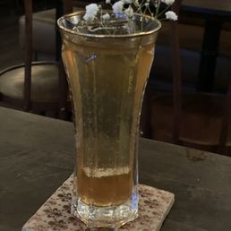 Fire Escape cocktail! - A tall, clear glass filled with a golden, bubbly drink, garnished with small white flowers, sits on a decorative coaster on a wooden table.