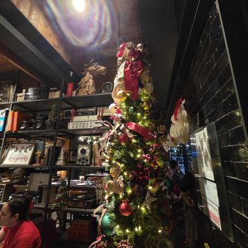 Haywire - A Christmas tree decorated with various ornaments, ribbons, and lights is positioned inside a restaurant. The background shows shelves with books, audio equipment, and decorative items, along with a black tiled wall and a wooden ceiling. There are people seated at tables, and festive decorations are visible, creating a holiday atmosphere. - New American Near Me - Austin, Texas
