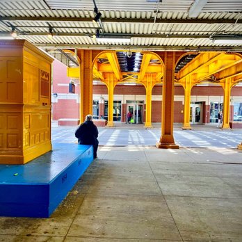 CTA - Diversey - A person is seated on a blue bench at a train station platform, with yellow structural supports and a brick building in the background. - Public Transportation Near Me - Chicago, Illinois