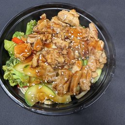 Hibachi Bowls - A takeout-style chicken hibachi bowl contains grilled chicken pieces topped with sauce and sesame seeds. The bowl also includes broccoli, zucchini, carrots, and other vegetables, arranged on a bed of rice or noodles. The dish is served in a black plastic container placed on a dark surface.