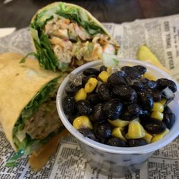 Baja Chicken wrap with Black Bean & Corn salad - A burrito and a wrap, both filled with lettuce and other ingredients, are placed on newspaper-lined paper, alongside a cup of black bean and corn salad.