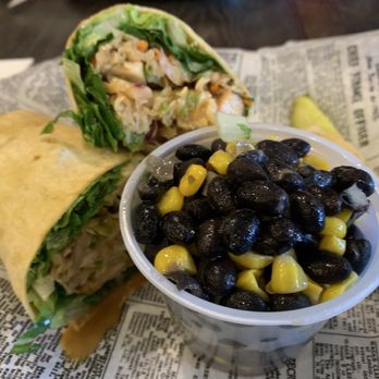 Lola's Cafe - Baja Chicken wrap with Black Bean & Corn salad - A burrito and a wrap, both filled with lettuce and other ingredients, are placed on newspaper-lined paper, alongside a cup of black bean and corn salad. - New American Near Me - Poughkeepsie, New York