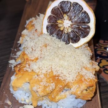 The 698 - A sushi roll is presented on a wooden serving board, topped with melted cheese and breadcrumbs. A dried lemon slice is placed on top as a garnish. The sushi roll appears to have a rice base with a flavorful topping, and the presentation includes scattered breadcrumbs around the roll. - Sushi Bars Near Me - Columbus, Ohio