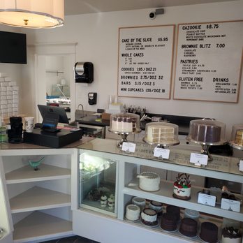 The Chocolate - Order Counter - The image depicts a dessert cafe counter with a glass display case containing assorted cakes, cupcakes, and decorated desserts. Behind the counter, there are menu boards listing prices for items such as cakes, cookies, brownies, and bars. The cafe has a modern interior with a white counter, a point-of-sale system, and a wall-mounted paper towel dispenser. The setting appears clean and organized, with a focus on baked goods and sweet treats. - Desserts Near Me - Orem, Utah