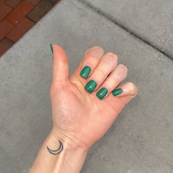 Ruby Nails - $45 for a full set with regular polish - The image features a person's left hand with neatly painted green nails, positioned against a concrete sidewalk. A small crescent moon tattoo is visible on the wrist. The person is wearing a black sleeve, and part of their red pants and black shoes can be seen at the bottom of the image. The background includes a concrete sidewalk and a section of brick pavement. - Nail Salons Near Me - Fairport, New York