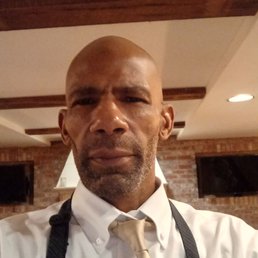 Me, before I showed up... - A man with a shaved head and beard, wearing a white dress shirt and beige tie, is taking a selfie. The background features brick walls, wooden ceiling beams, and two wall-mounted TV screens, indicating an indoor environment such as a restaurant or bar.