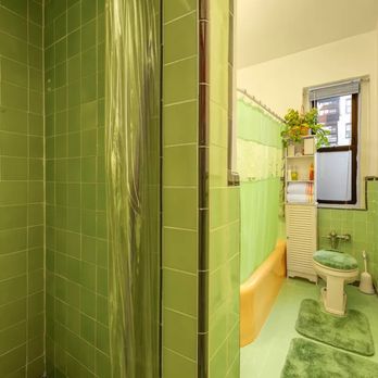 Build Pro - Before - A compact bathroom features green tiled walls, a shower area with a metal showerhead, and a toilet with a green seat cover. A window with a plant and storage shelf is visible. - General Contractors Near Me - West Babylon, New York