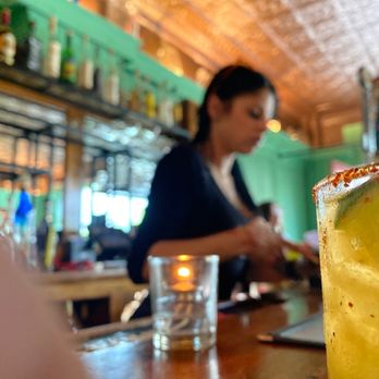 Armadillo - Infinity - A margarita with a salted rim and lime wedge is in focus on a bar counter, with a blurred bartender in the background. - Mexican Near Me - Kingston, New York
